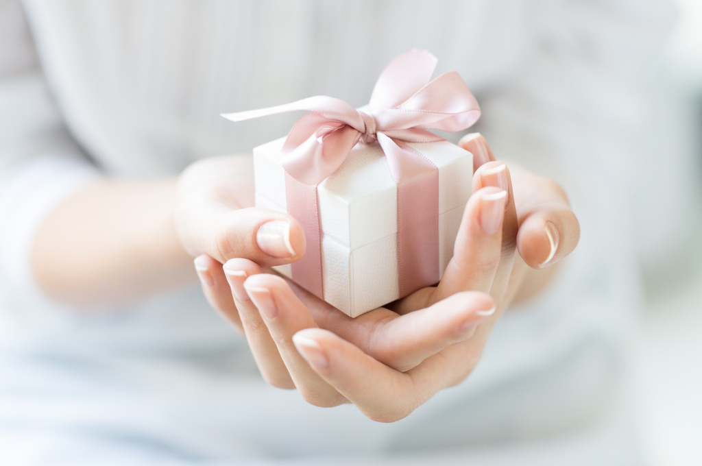 Close up shot of female hands holding a small gift wrapped with pink ribbon. Small gift in the hands of a woman indoor. Shallow depth of field with focus on the little box.